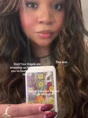 A close-up shot featuring a person's face and hands. The person has long, wavy brown hair and is holding a deck of tarot cards. Text overlays on the image read, 'God/Your Angels are wrapping up the you've been QUICK', and 'The end'. Below, a smaller text says, 'Do they know that they got 30 minutes?'. The person's face fills most of the frame, and their eyes are looking directly at the viewer. Their nails are painted with a deep red color, and they wear a ring. The background is blurred, giving focus to the subject and the card.