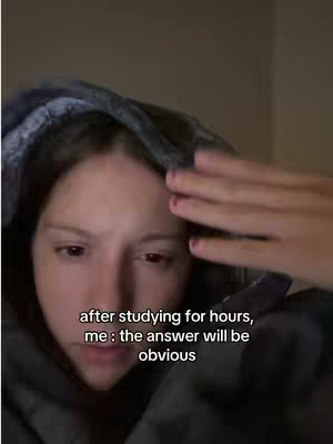 A close-up shot of a person's face, partially obscured by a dark hoodie. Text overlayed on the image reads "after studying for hours, me : the answer will be obvious". The person has fair skin, dark hair partially visible under the hood, and red nail polish. They are looking down and to the left, and have a hand raised near their head.