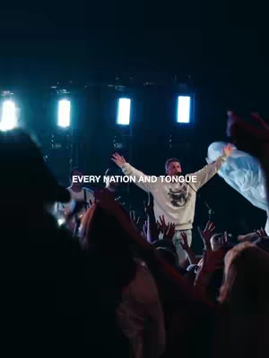 The image captures a live performance with a man in the center, arms raised, seemingly leading worship or giving a speech. Soft lighting from the background adds to the atmosphere. The crowd, slightly blurred, extends towards the bottom of the frame, with arms raised, emphasizing the communal and emotional nature of the event. Text overlayed indicates the key phrase.