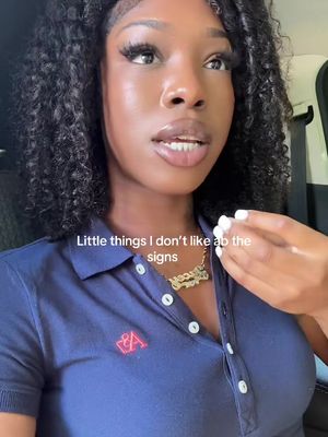 A woman is shown in a selfie-style shot, angled slightly up from her chest. She has curly black hair and is wearing a navy blue polo shirt with a small logo on the left chest. A gold necklace with the name "Marissa" is around her neck. The background is slightly blurred, with a hint of a vehicle interior visible. The lighting seems natural, and the overall composition is simple and focused.