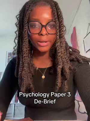 A selfie-style shot of a young woman wearing glasses and a black top is presented. She's centered in the frame, seemingly looking directly at the camera, with a soft, focused expression. Her hair is styled in long, thick braids. The background is somewhat blurred, with hints of an interior space and wall art. The text overlay at the bottom states 'Psychology Paper 3 De-Brief'.