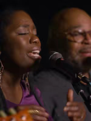 The cover image shows two people singing with microphones. The image has a low light setting, with a dark background. The person on the left is a Black woman looking upward with her eyes closed while singing into a microphone. She is wearing a purple top and has earrings. To the right is a blurred man with a black coat and glasses supporting the song. The shot appears to be a concert experience.
