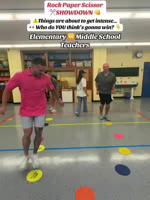 The cover image shows a group of people in a classroom setting, likely a gym or multi-purpose room. The image is a medium shot, with four people. The background indicates a school environment, with cabinets, a whiteboard, and a bulletin board visible. The lighting appears to be natural, coming from overhead fixtures. In the foreground, two men are running, one to the left with a pink shirt and the other to the right with a green shirt. The flooring is grey and covered with spots on the ground. A girl with a white t-shirt, blue jeans and a dark cap is standing at a distance. The overall palette is a mix of the natural colors of the environment with bold and contrasting colors on the ground.