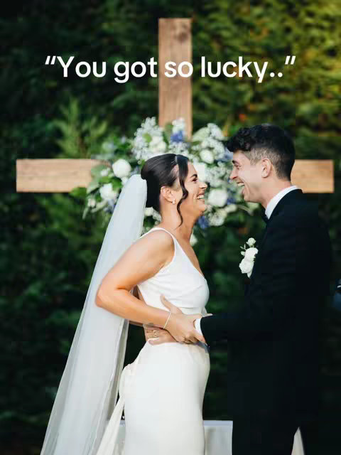 A wedding photo, featuring a smiling bride and groom as they embrace in front of a wooden cross decorated with white and blue flowers. The bride wears a white wedding dress and veil, the groom in a black suit. The background has lush green foliage.