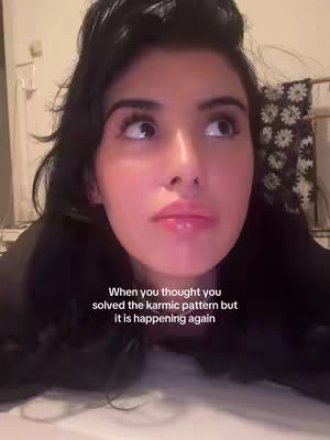 A close-up shot of a woman's face, looking slightly upward with a contemplative expression. Her dark hair is styled away from her face, and she has visible eye makeup and glossy lips. The background is blurred, suggesting a domestic setting. Text overlaid on the image states, 'When you thought you solved the karmic pattern but it is happening again'.