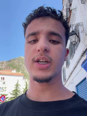 A young man with short, dark, curly hair and a slight beard is centered in the frame, facing forward and speaking. He wears a black t-shirt. Behind him is a bright blue sky and white buildings with blue accents, suggesting a Mediterranean or Moroccan town setting. To the left, a green hill is visible. The lighting is bright, indicating daytime.