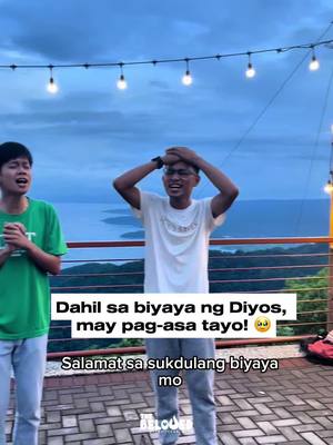 Two young men stand outdoors with a scenic background of a lake and cloudy sky. The man on the right has his hands on his head, smiling, with glasses. The man on the left is also looking at the camera, singing. Overlaid text in a white box reads, "Dahil sa biyaya ng Diyos, may pag-asa tayo!" (Because of God's grace, we have hope!) and "Salamat sa sukdulang biyaya mo" (Thank you for your great grace).