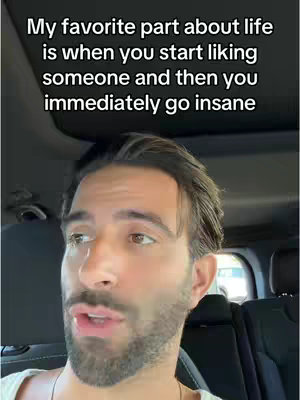 A man is filmed from the chest up, inside a car with the driver's seat and the roof visible. The man has short dark hair, a beard, and is facing the camera. Text on the top of the screen states: “My favorite part about life is when you start liking someone and then you immediately go insane.” 