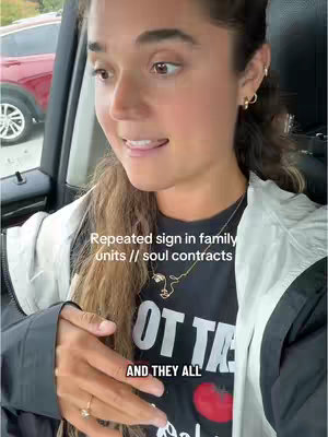 The cover image is a selfie-style shot taken from inside a car. A light-skinned woman with dark hair is the main subject, positioned slightly off-center. She looks toward the left of the frame with her mouth open, as if she is speaking. Her hair is styled with loose waves. She wears a necklace, earrings, and a black t-shirt with a graphic design on it. The background is out of focus, but it appears to be a car interior, with glimpses of the exterior showing, including a red car. A text overlay is present, reading "Repeated sign in family units // soul contracts". The overall composition is casual and informative.