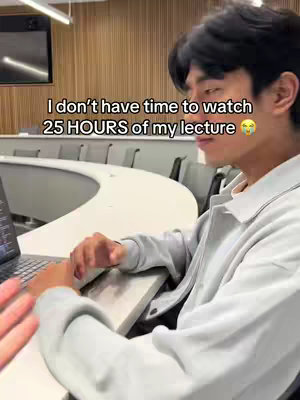 A young man is sitting at a desk next to a laptop in what appears to be a classroom. He is looking at the laptop and a text overlay on the image says, 'I don't have time to watch 25 HOURS of my lecture' with a surprised emoji.
