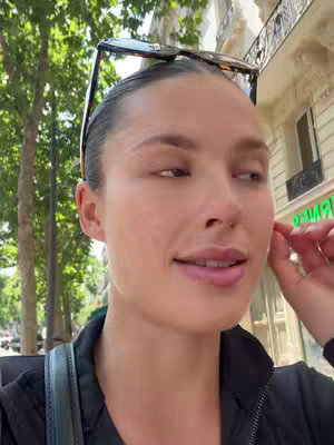 A woman is walking on a city street, with buildings and trees in the background. She has sunglasses perched on her head and is adjusting a strand of hair. The lighting suggests a sunny day.
