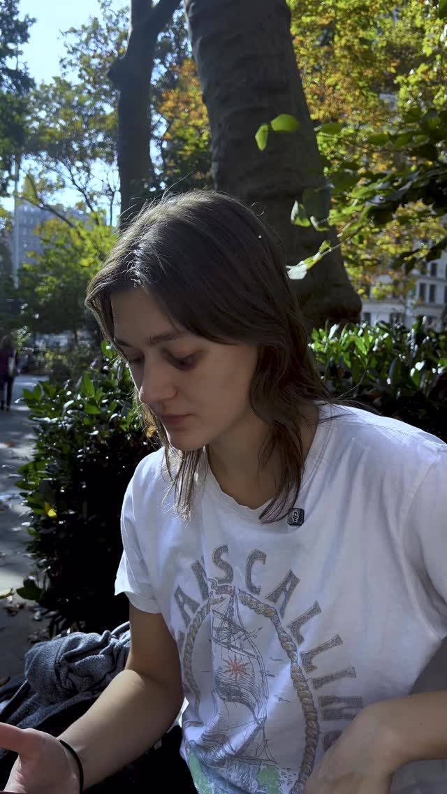 A woman with brown hair is seated outdoors, facing downwards with a pensive expression. She is wearing a white t-shirt with a graphic design and a small microphone is visible near her face. The background shows trees with green and yellow leaves, a building, and filtered sunlight.