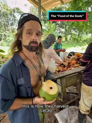 A man holding a partially cut green cacao fruit stands in front of a table with several people. Text overlays state "The Food of the Gods".