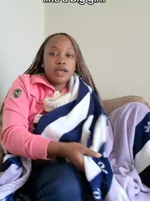 A woman is sitting on a sofa, wrapped in a striped blanket wearing a pink jacket. The blanket is navy blue and white, and a lilac blanket is also visible. The woman is looking towards the camera with a neutral expression.