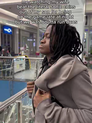 A man with dreadlocks wearing a gray hoodie stands in a mall setting, looking off to the side with a slightly pensive expression. Overlaid text describes a humorous situation involving his family and video games.