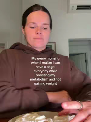 A woman in a brown hoodie is shown from the chest up, centered in the frame. She is looking down slightly, positioned in what appears to be a kitchen or dining space. A plate of food, likely bagels with topping, is visible in the foreground, her hand is on the plate. Text overlay is present, describing the image.