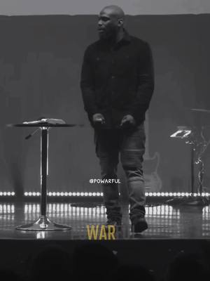 A black and white image showing a man speaking on stage with a microphone stand and a table in front of him. The word 'WAR' is overlaid on the bottom.