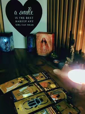 The cover image shows a low-angle shot of a table with tarot cards fanned out. In the background, there is a wooden wall and a heart-shaped art piece that reads, "a smile is the best makeup any girl can wear." Various spiritual decorations, including lit candles and a statue, complement the arrangement, and a deck of cards is visible.