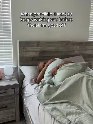 A person is lying in a bed, mostly covered by a blanket and holding a pillow to their face. The room is brightly lit, with visible blinds on the window. A nightstand with a lamp is on the left.