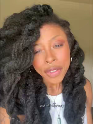 A woman with long, dark, curly hair is looking towards the camera. She has a nose ring, and is wearing a white top. A pendant hangs from a necklace.