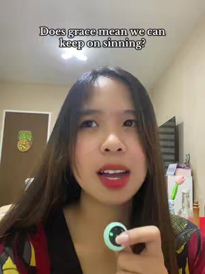 A woman with long dark hair is in front of a blurry indoor background. She is looking up and to the left. The text overlay is in a black box at the top of the frame, the text says Does grace mean we can keep on sinning?. She is holding a small, round microphone in her right hand in front of her chest.