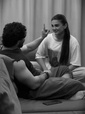 A black and white photo of a couple sitting close together. A man with dark curly hair is leaning back on a couch, with his arm extended. A woman with long hair is sat next to him, smiling and pointing upwards. The background is simple, indicating an indoor setting.