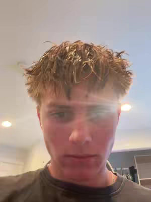 A close-up shot of a young man's face, with light-colored hair and a neutral expression. The background is a blurred, bright interior.