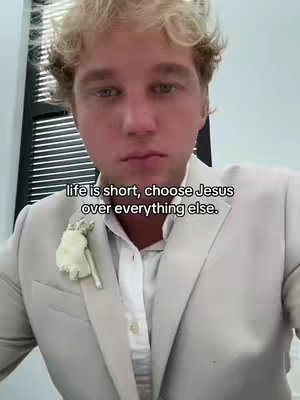 A close-up of a young man wearing a light-colored suit jacket, with text overlayed. He has short, curly blonde hair and is looking towards the camera. A white flower is pinned to his lapel. The background is out of focus, with a glimpse of a window and some dark blinds visible.