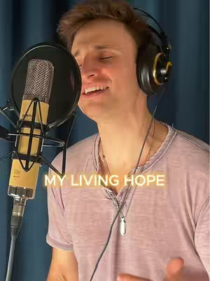 A man with short brown hair is singing into a microphone. He's wearing a light pink shirt and headphones. Overlayed in yellow text is the phrase "MY LIVING HOPE". The background appears to be a dark blue curtain.