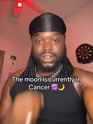 A man with dreadlocks wearing a black durag and tank top speaks directly to the camera. White text overlayed on the image reads, \"The moon is currently in Cancer ♋️🌙\". The background is a room with a red ceiling fan, a dartboard, and a curtain.