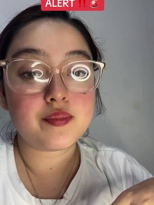 A close-up shot featuring a woman with fair skin, wearing large, clear-framed glasses. Her hair is pulled back, and she's wearing a white top. A red banner with the word 'ALERT' is at the top of the image. The background is a plain gray. The woman's lips are painted with a red lip color and she has a gold necklace.