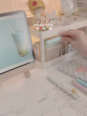A softly lit photo of a study desk with an open iPad displaying a drink and straw. A small shelf holds cute bunny figurines and other items. Handwriting and notes are visible on the desk surface with a pen laid down, suggesting study materials.