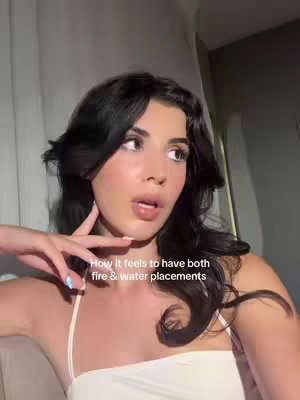 A woman with dark hair looks upwards with a slight open mouth expression in front of a light background. Text overlayed on the image reads, "How it feels to have both fire & water placements".