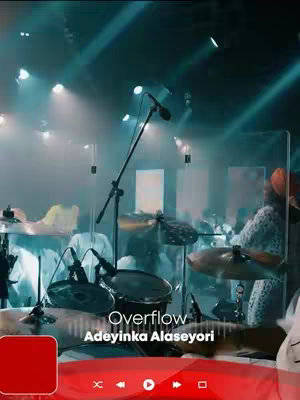 The cover image captures a dynamic live music performance scene. The foreground features a drum set, with cymbals and drums clearly visible. In the background, a stage is set up with lighting highlighting a band performing. A text overlay at the bottom announces the title and artist. The overall composition suggests a concert or music video setting, with cool tones of blue.