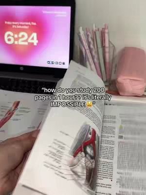 A person is holding an open book with anatomical diagrams, with a laptop displaying a pink-colored timer on the screen. The book is close to the camera, and the background includes a clear container of pink pens, and a pink item, possibly a container or organizer.