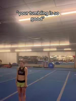 A woman in a cheerleading uniform stands on a blue mat in a gymnasium, with text overlayed on the image. The image suggests a focus on cheer or tumbling.
