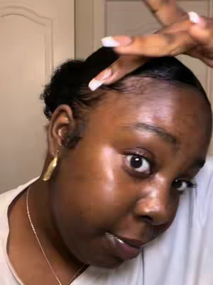 A close-up shot of a woman's face, angled slightly. She has dark skin, is wearing gold earrings and a gold necklace. Her hair is styled with her baby hairs brushed down. She has light-colored, manicured, square-shaped nails. The background is blurred, likely a neutral wall. The lighting is soft, highlighting her features, particularly her skin's luminosity, and the edges of her hair and edges.