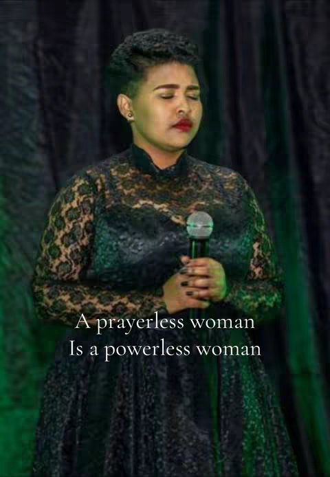 A woman with dark skin stands in a dimly lit room. She wears a black, intricately designed gown with sleeves and a high neckline. Her hair is styled in a short, textured updo, and she has a serious expression, with her eyes closed and her lips slightly parted. She holds a microphone with both hands, as if speaking or singing. Overlayed text reads, 'A prayerless woman is a powerless woman.' The background is a dark green fabric
