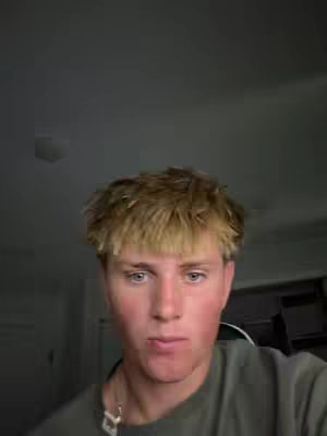 A young man with light brown hair looks directly at the camera. He is wearing a green t-shirt or top and a silver necklace. The background is a dimly lit interior.