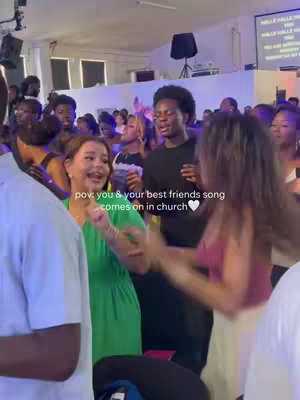 The image captures a lively scene inside a church, filled with a crowd of people. The focus of the image is on a woman in a green dress who appears to be singing and enjoying herself. Next to her a man in a black shirt is singing along. The background shows other people, many of whom are also singing and raising their hands. Above them, a screen displays words, giving the impression of a worship or service setting. The overall vibe conveys a sense of community, spiritual engagement, and praise.