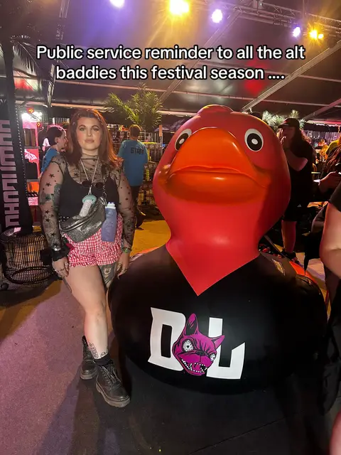 A woman stands with a large inflatable red duck wearing a black t-shirt with the Download Festival logo. Text overlayed says "Public service reminder to all the alt baddies this festival season ...." The woman is dressed in a black top, pink shorts, and boots.