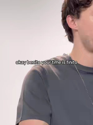 A medium shot shows a person from the chest up. The person is facing slightly towards the camera with the face angled slightly to the side. The person wears a gray t-shirt and has a silver chain around their neck. overlaid on the image is the white text: okay benito your time is finito