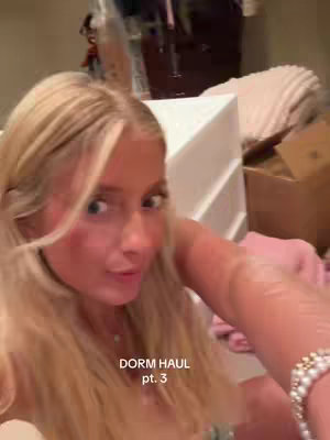 A selfie-style video cover, featuring a young woman with blonde hair and fair skin. She is positioned in the foreground, looking towards the camera with a slight smile. The background is slightly blurred, showing what looks like a dorm room with boxes and furniture. Text overlayed on the bottom of the image reads "DORM HAUL pt. 3". The overall tone suggests a vlog or haul video.
