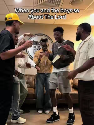 A group of young Black men are gathered in what looks like a lounge or studio setting. One man in a yellow baseball cap stands at the left edge, while several others are in the frame, some with arms raised and a mix of expressions. The background includes a round sign with text, a tan sofa, and overall warm lighting.