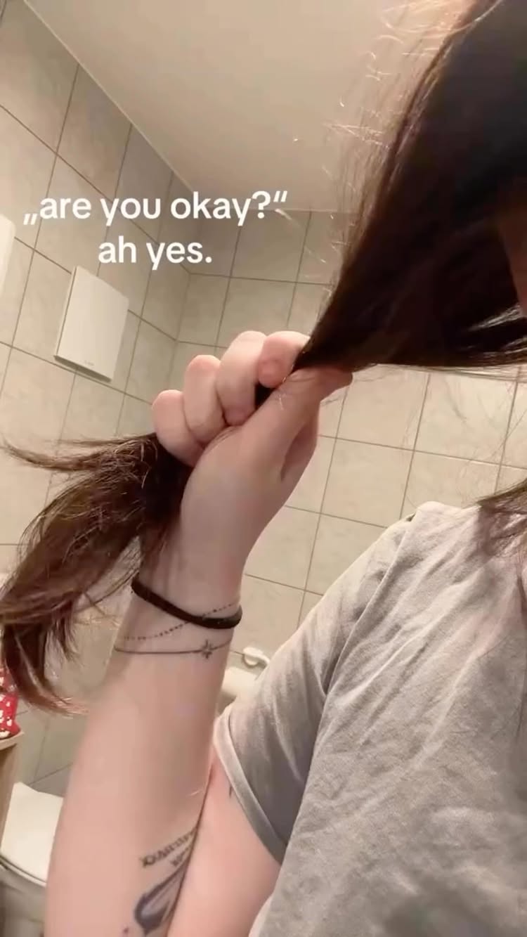 The cover image shows a close-up of a person's hand holding a large section of brown hair. The person is wearing a gray t-shirt and has a tattoo with the word "XANADU" on their arm. The background is a bathroom with white tiles. The text overlay says "are you okay?" ah yes.