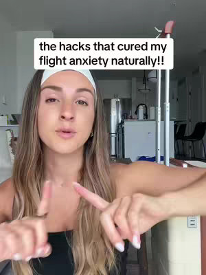 A woman with long, light brown hair is shown from the chest up, gesturing with her right index finger while pointing with her left index finger. A white text overlay is at the top of the frame. She's wearing a black tank top and a white headband. The background includes a glimpse of an apartment setting with a glimpse of a kitchen and a metal stand.