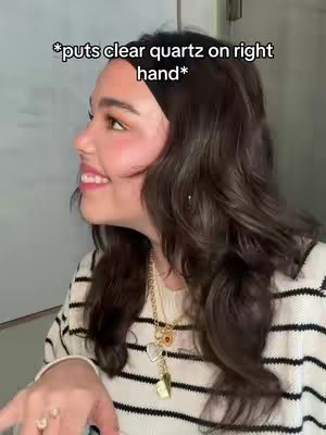 A woman with long, dark wavy hair is seen in a side profile, looking slightly to the upper right of the frame. She has rosy cheeks and a subtle smile, possibly applying makeup. She wears a black-and-white striped sweater and multiple necklaces which add a nice accent. The background appears to be a neutral gray, and overlaid text is present, indicating a focus on clear quartz and its spiritual properties.