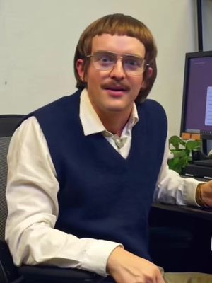 A man with a bowl haircut, glasses, and a mustache is sitting at a desk. He is wearing a white collared shirt under a dark blue vest. There is a computer monitor to his right and a dark desk in front of him.