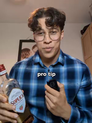A young man with curly brown hair, glasses, and a blue plaid shirt smiles while holding a bottle of liquor in one hand and a phone in the other. A text overlay says 'pro tip'.