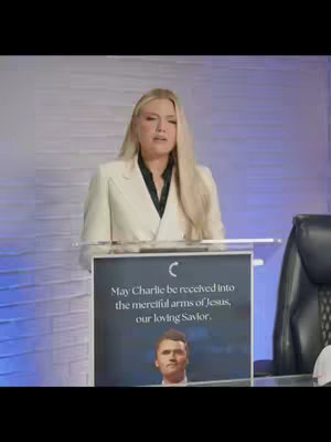 A woman in a white blazer stands behind a clear podium. Behind her is a light blue wall with a textured pattern. The podium has a sign with a picture of a man and text. To the woman's right is a black chair.